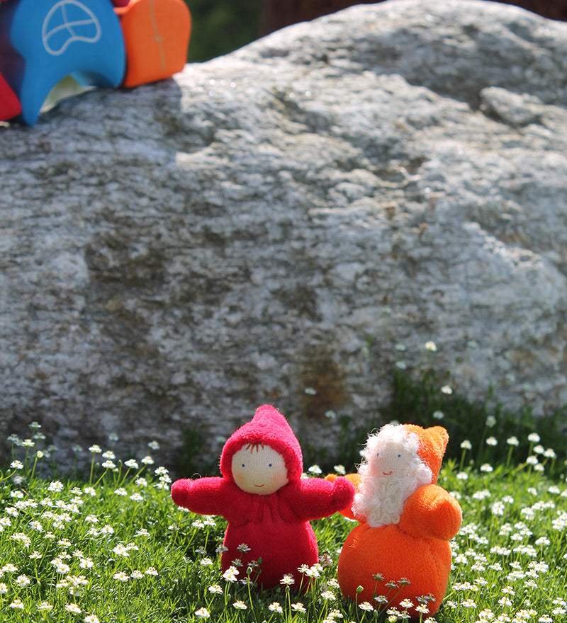 An orange and red Grimm's Dwarf doll enjoying the sunshine and stood on a patch of grass full of small daisies. Behind the dwarf toys is a large grey rock and a Grimm's House