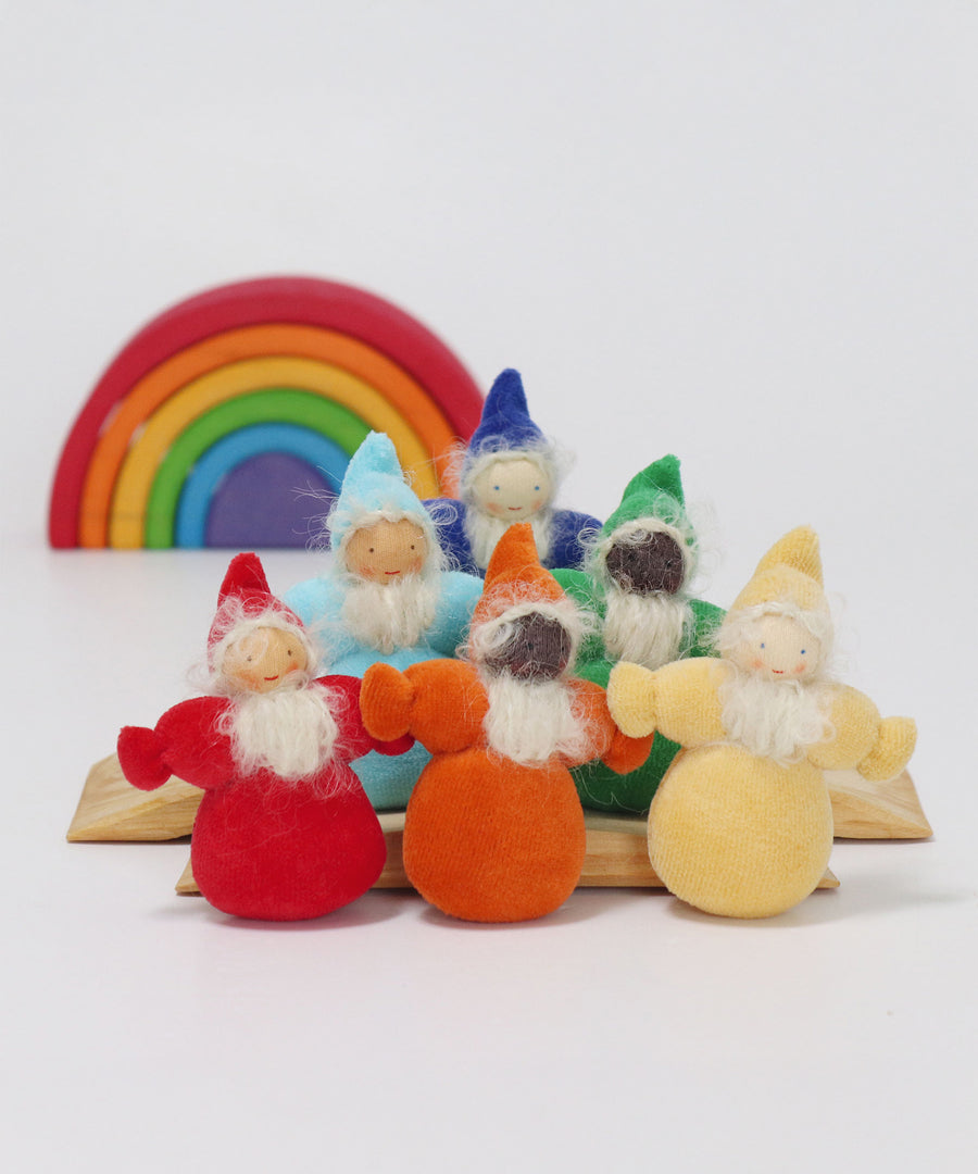 The Grimm's Rainbow Dwarfs collection showing a red, orange, yellow, light blue, green and indigo Grimm's Rainbow Dwarfs on natural wood blocks, with a mini Grimm's rainbow in the background