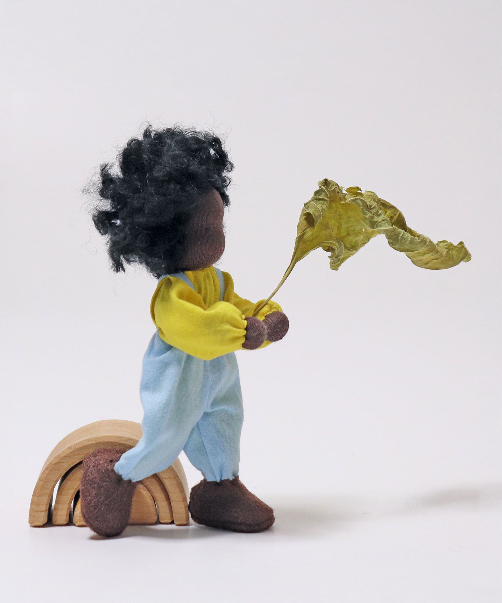 Grimm's Handmade Doll - Max is stood next to a Grimm's small natural rainbow and is holding a green leaf in its hand. The image is on a grey background