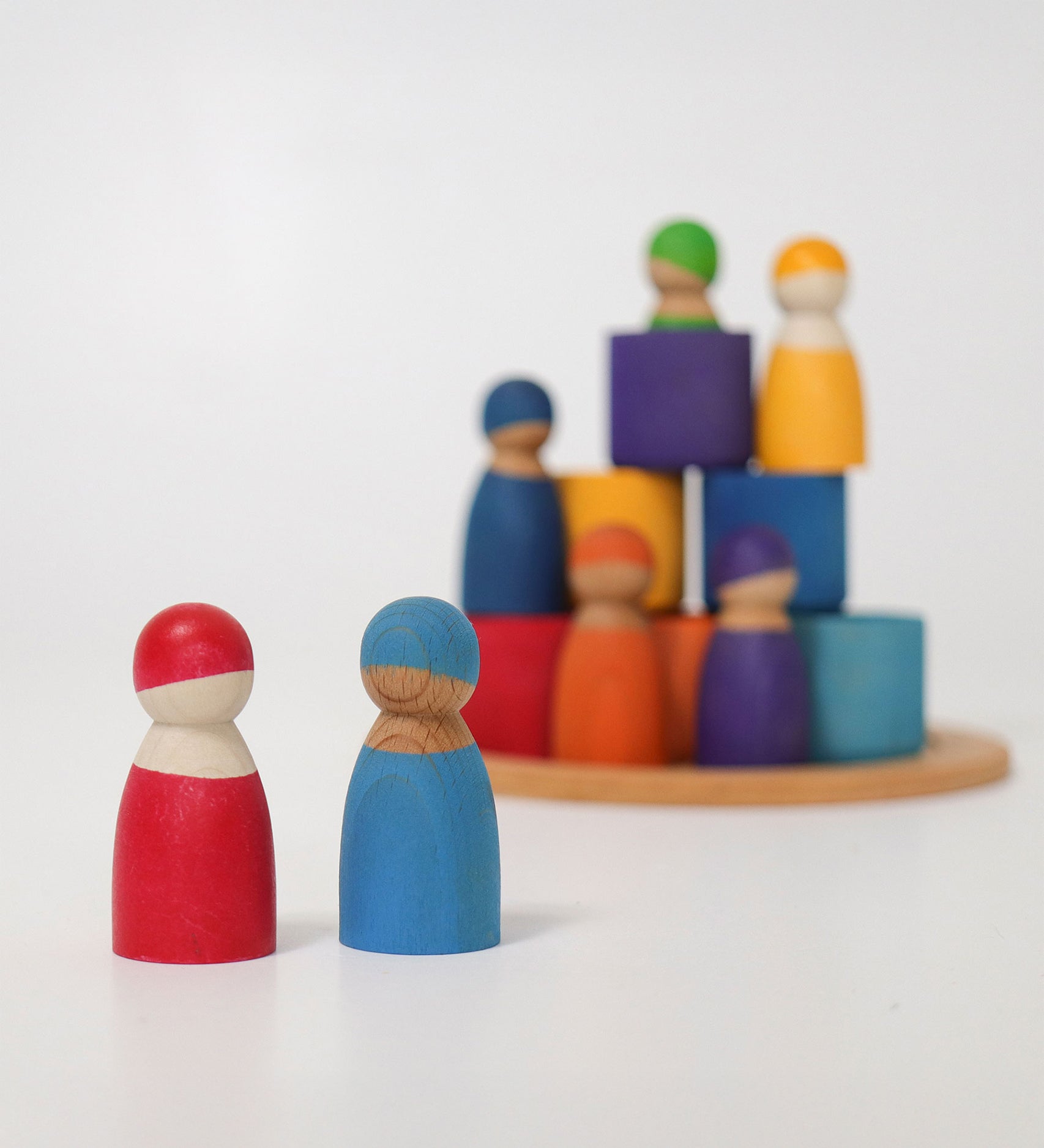 Two Grimm's Rainbow dolls in the foreground with the Wooden Rainbow Bowls and other Grimm's Rainbow Dolls stacked in the background. The focus is on the two peg dolls at the front with the pieces in the background blurred slightly