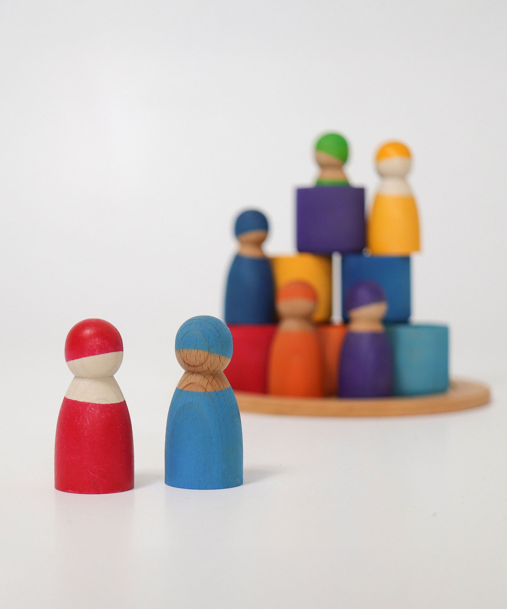 Two Grimm's Rainbow dolls in the foreground with the Wooden Rainbow Bowls and other Grimm's Rainbow Dolls stacked in the background. The focus is on the two peg dolls at the front with the pieces in the background blurred slightly