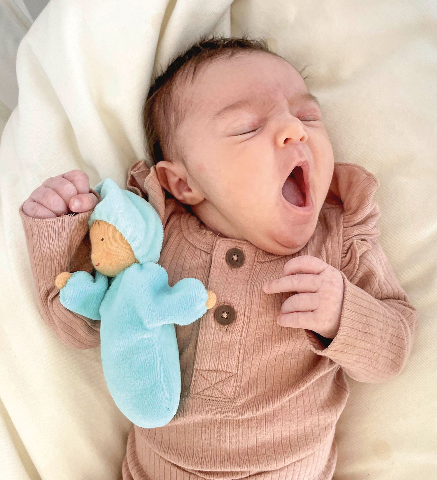 A small baby is yawning whilst sleeping, and is cuddles with Grimm's Lavender Doll Aqua