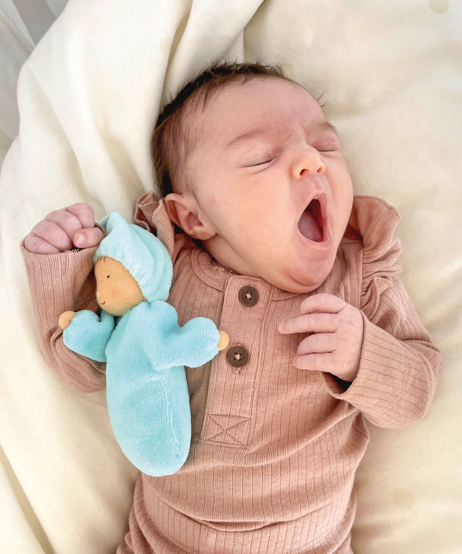 A small baby is yawning whilst sleeping, and is cuddles with Grimm's Lavender Doll Aqua