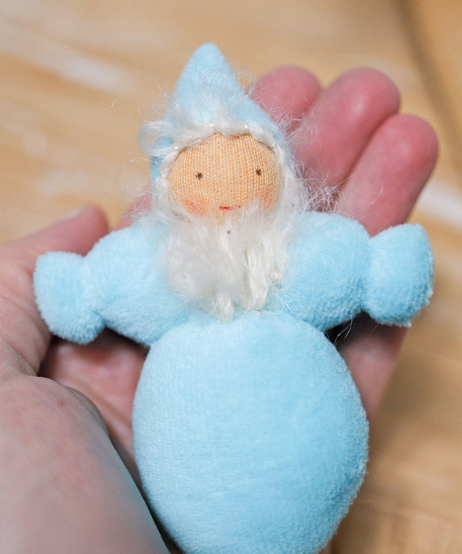 A closer view of the light blue Grimm's Rainbow Dwarfs, in a persons hand. The image shows the dwarfs body, bear, smiling face and hat up close and gives a size guide as it fits in the palm of an adults hand.