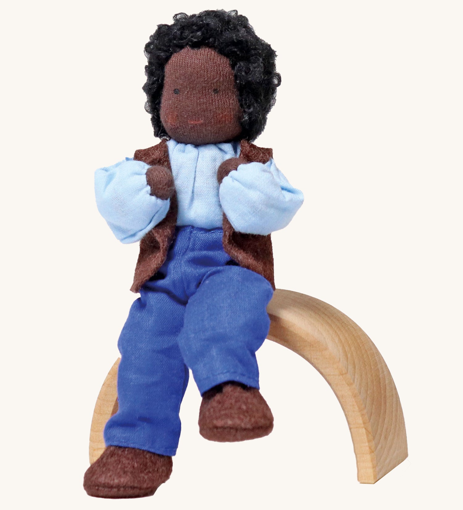 Grimm's Handmade Doll - Mr Ebony, is sitting on a natural rainbow arch. The doll has black skin, black curly hair, a smiling face, and is wearing brown fabric shoes, blue trousers, a blue top and a brown sleeveless jacket. The image is on a cream background.