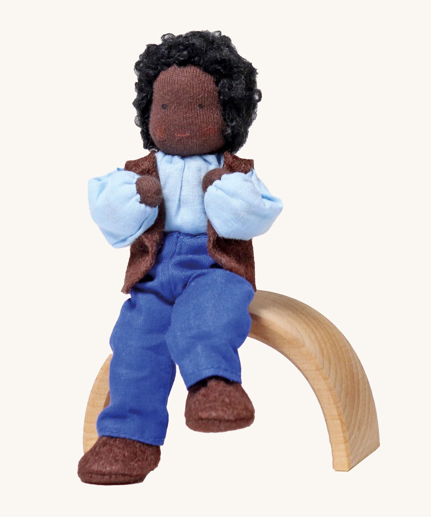 Grimm's Handmade Doll - Mr Ebony, is sitting on a natural rainbow arch. The doll has black skin, black curly hair, a smiling face, and is wearing brown fabric shoes, blue trousers, a blue top and a brown sleeveless jacket. The image is on a cream background.