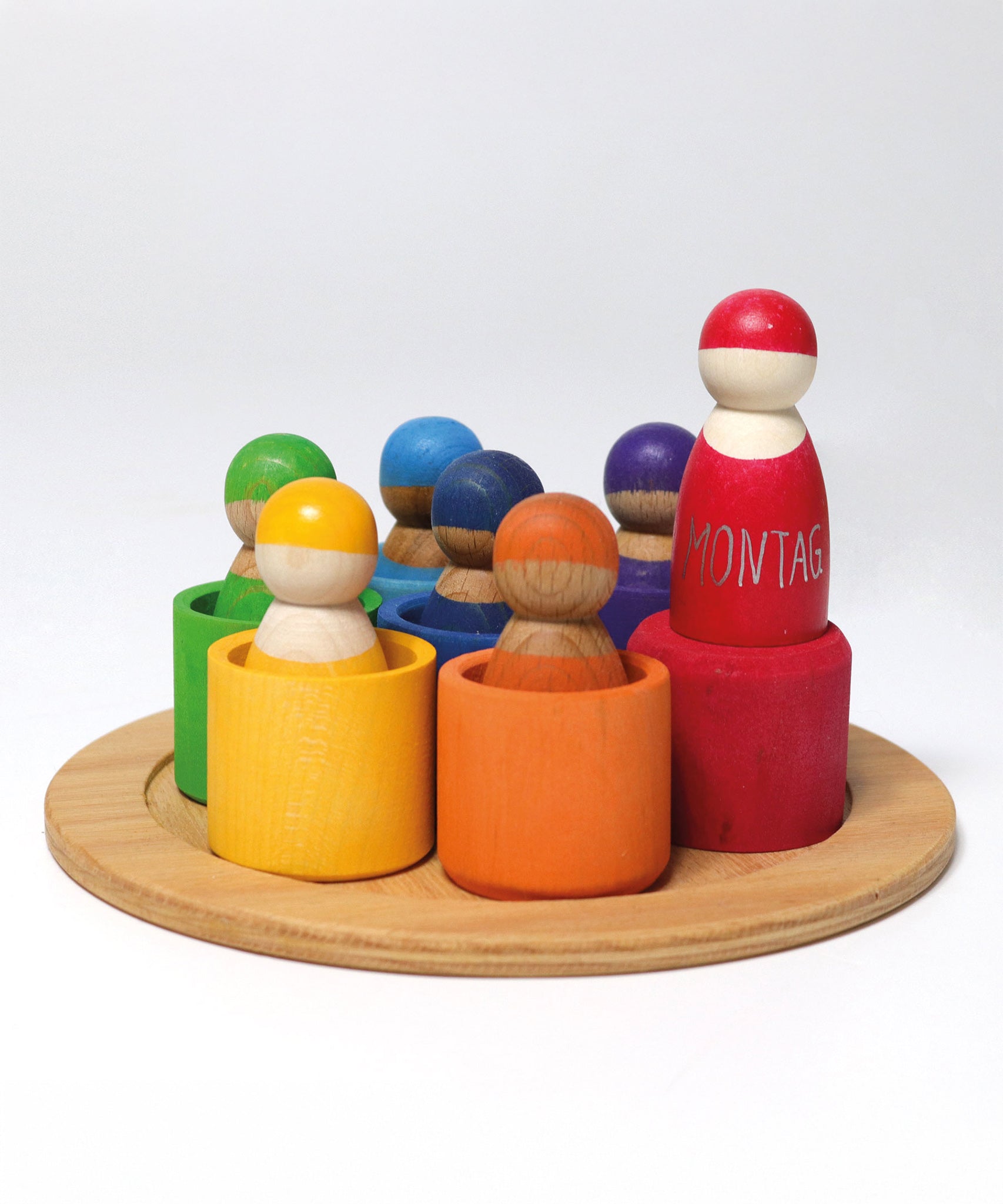 The Grimm's Wooden Rainbow Friends with bowls, stood on a small wooden plate on a cream background. The red bowl has been turned upside down and the red peg doll is stood on top 