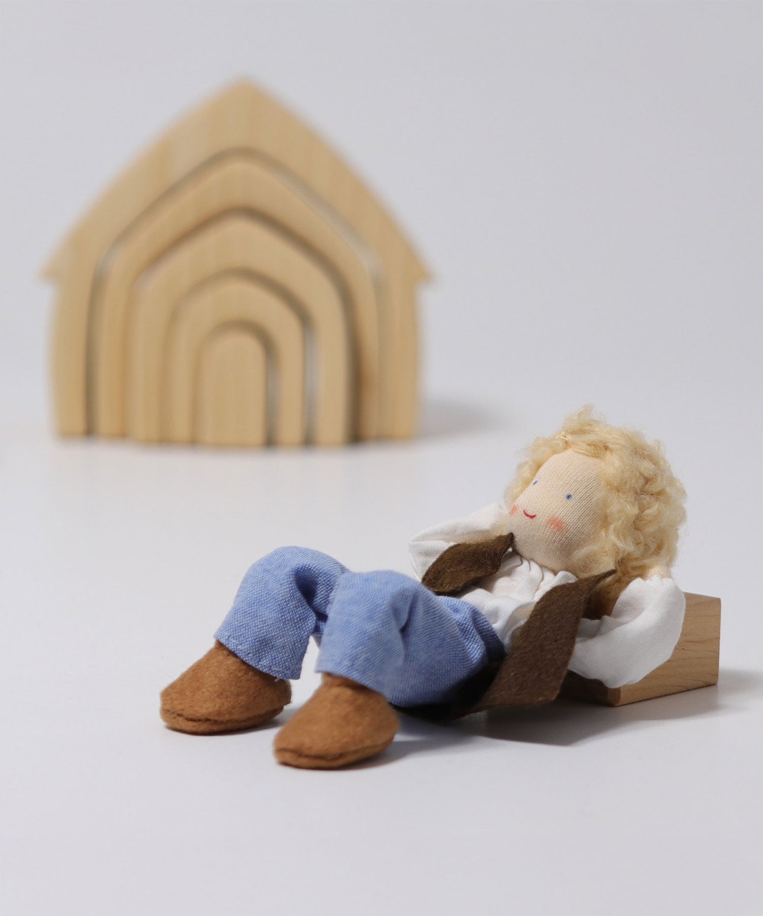 The Grimm's Blond Haired Man Doll, laying down on its back with its arms behind its head, and its knees bent in a relaxed position. There is a Grimm's Natural House in the background.