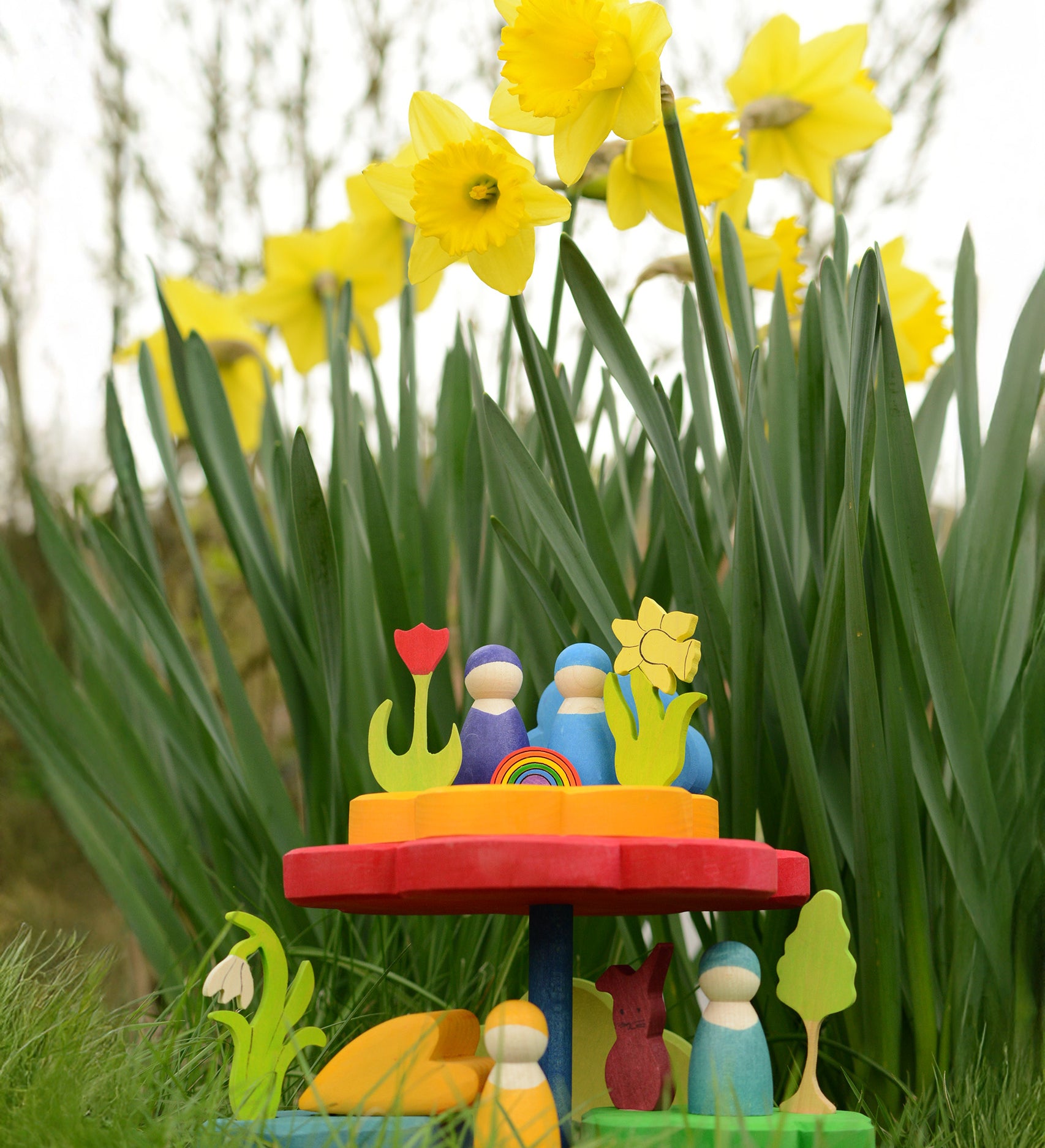 The Grimm's Stacking Flowers Tower, is set down on lush green grass, in front of a patch of daffodils. The stacker has been made into a little display with Grimm's decorative figures and Grimm's friends