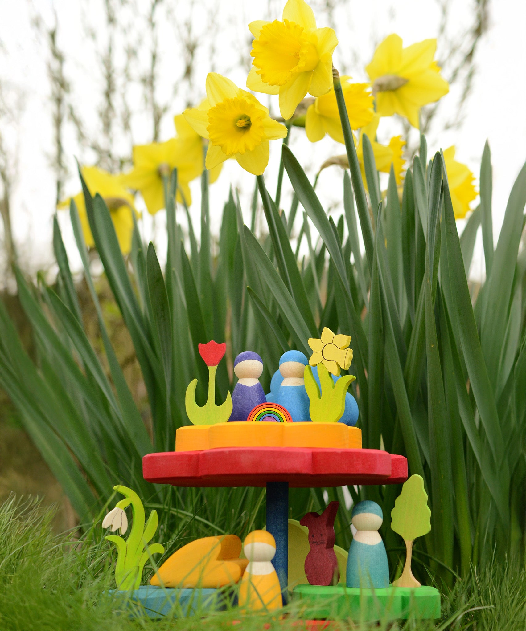 The Grimm's Stacking Flowers Tower, is set down on lush green grass, in front of a patch of daffodils. The stacker has been made into a little display with Grimm's decorative figures and Grimm's friends