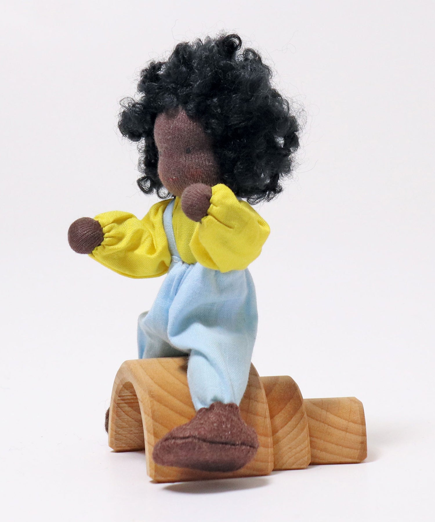 A side view of Grimm's Handmade Doll - Max sitting on a Grimm's small natural rainbow. The image is on a light grey background