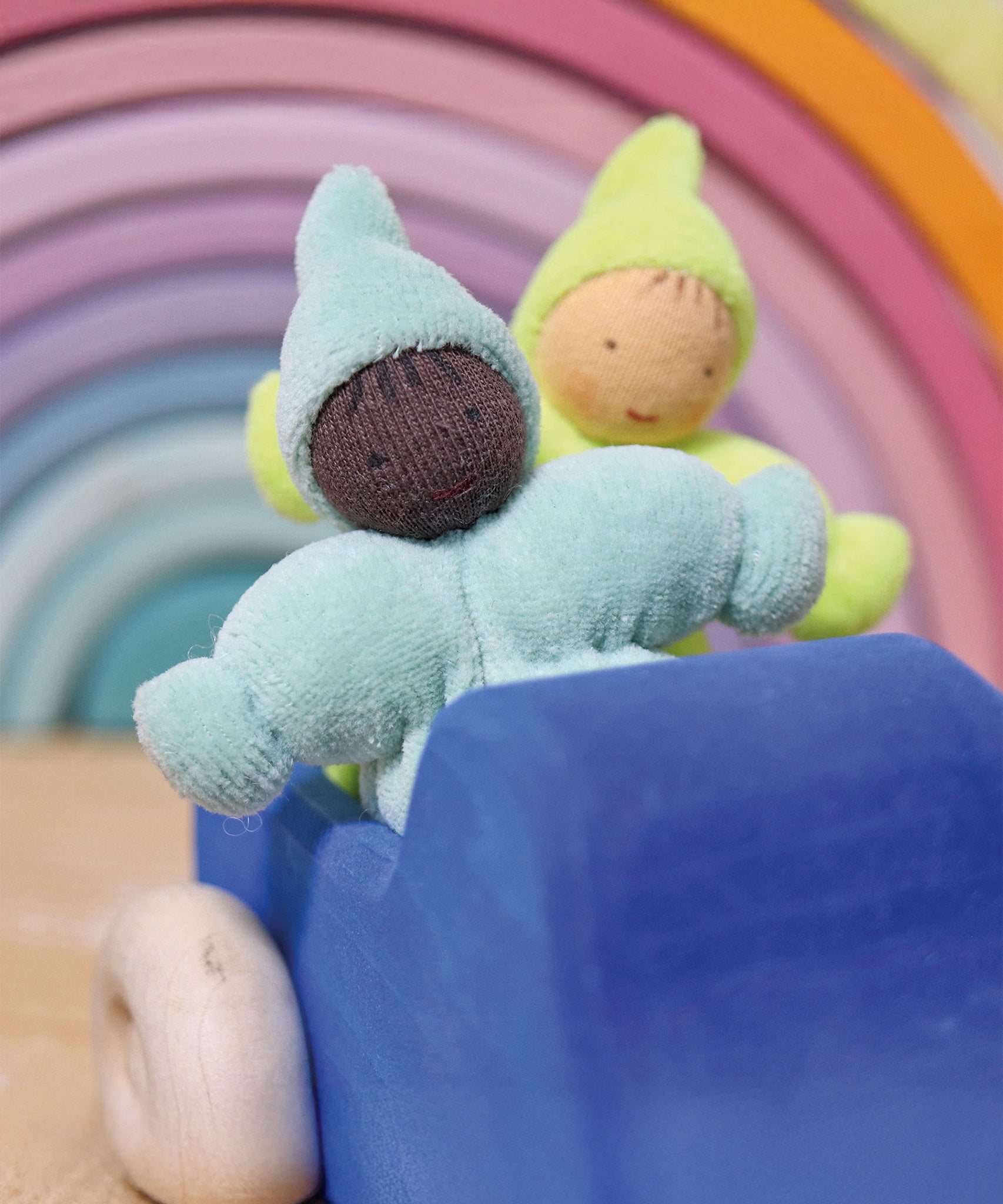 The pastel blue and pastel green The Grimm's Pastel Dwarfs dolls are sat in a wooden blue Grimm's car toy, with a Grimm's pastel rainbow in the background