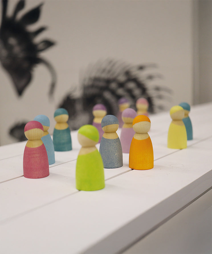 The Grimm's 12 Pastel Friends are all stood together on a white wooden table, with a black art silhouette in the background