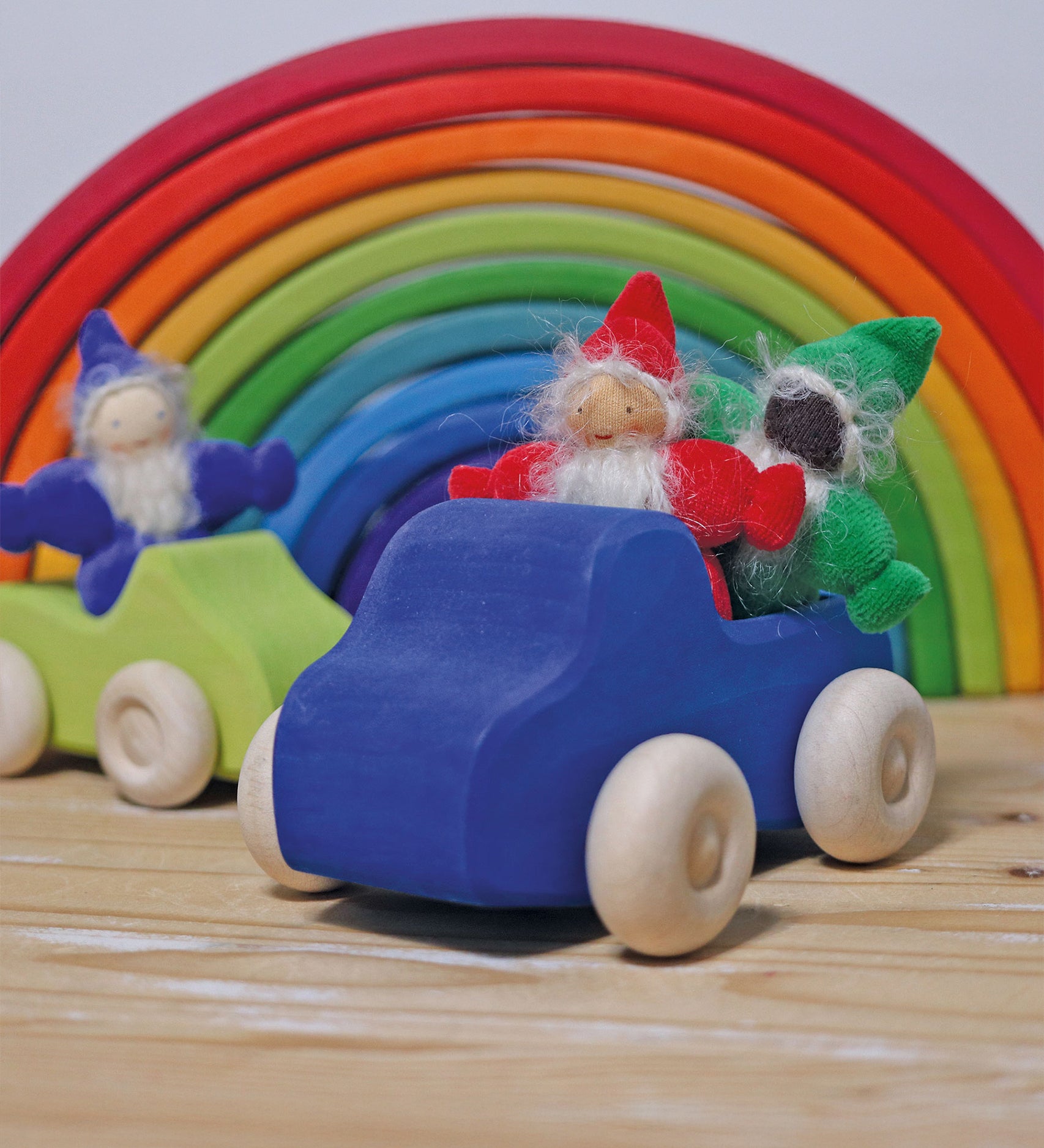 The red, green and indigo Grimm's Rainbow Dwarfs are sat in Grimm's wooden car toys with a large Grimm's rainbow in the background