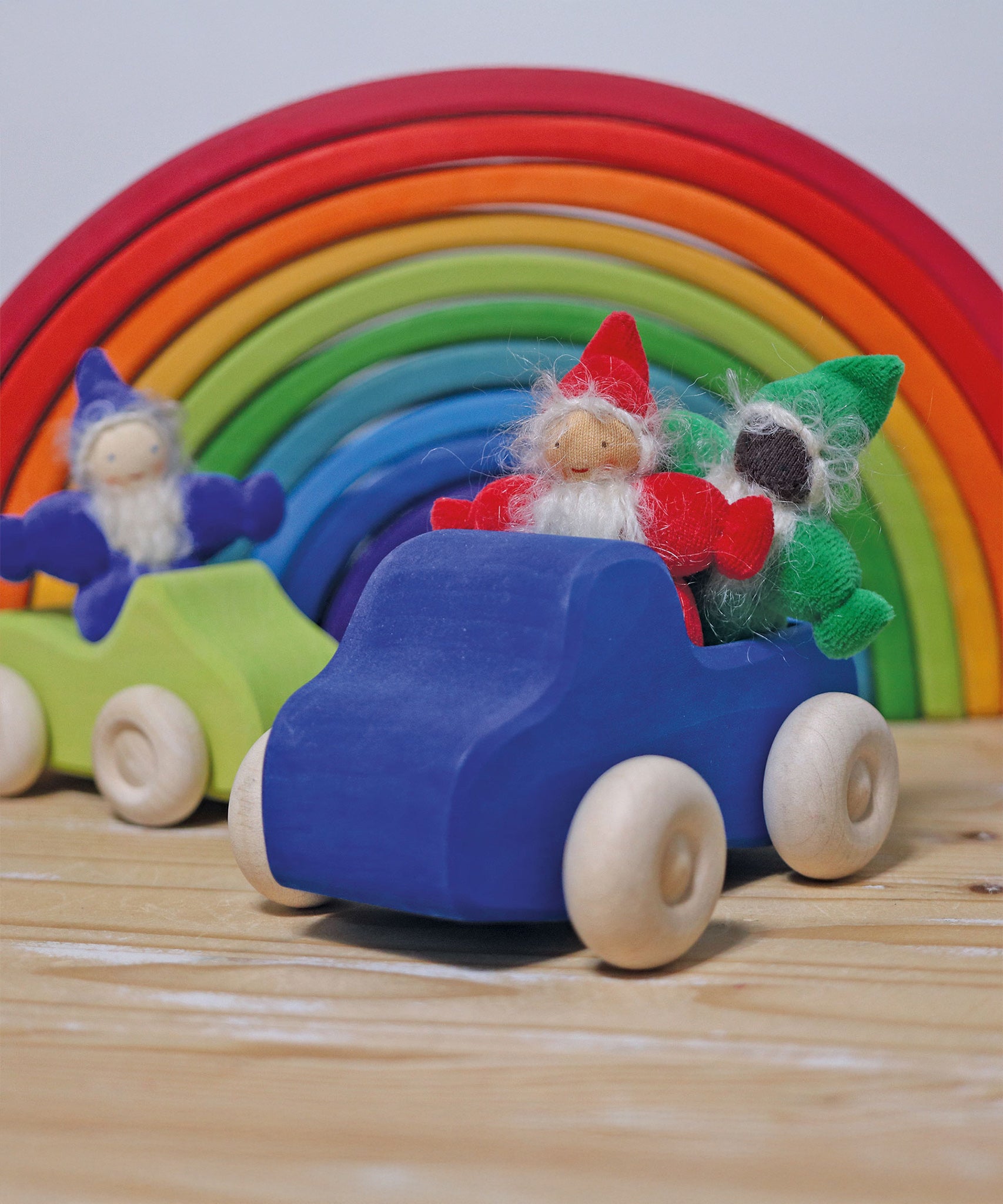 The red, green and indigo Grimm's Rainbow Dwarfs are sat in Grimm's wooden car toys with a large Grimm's rainbow in the background