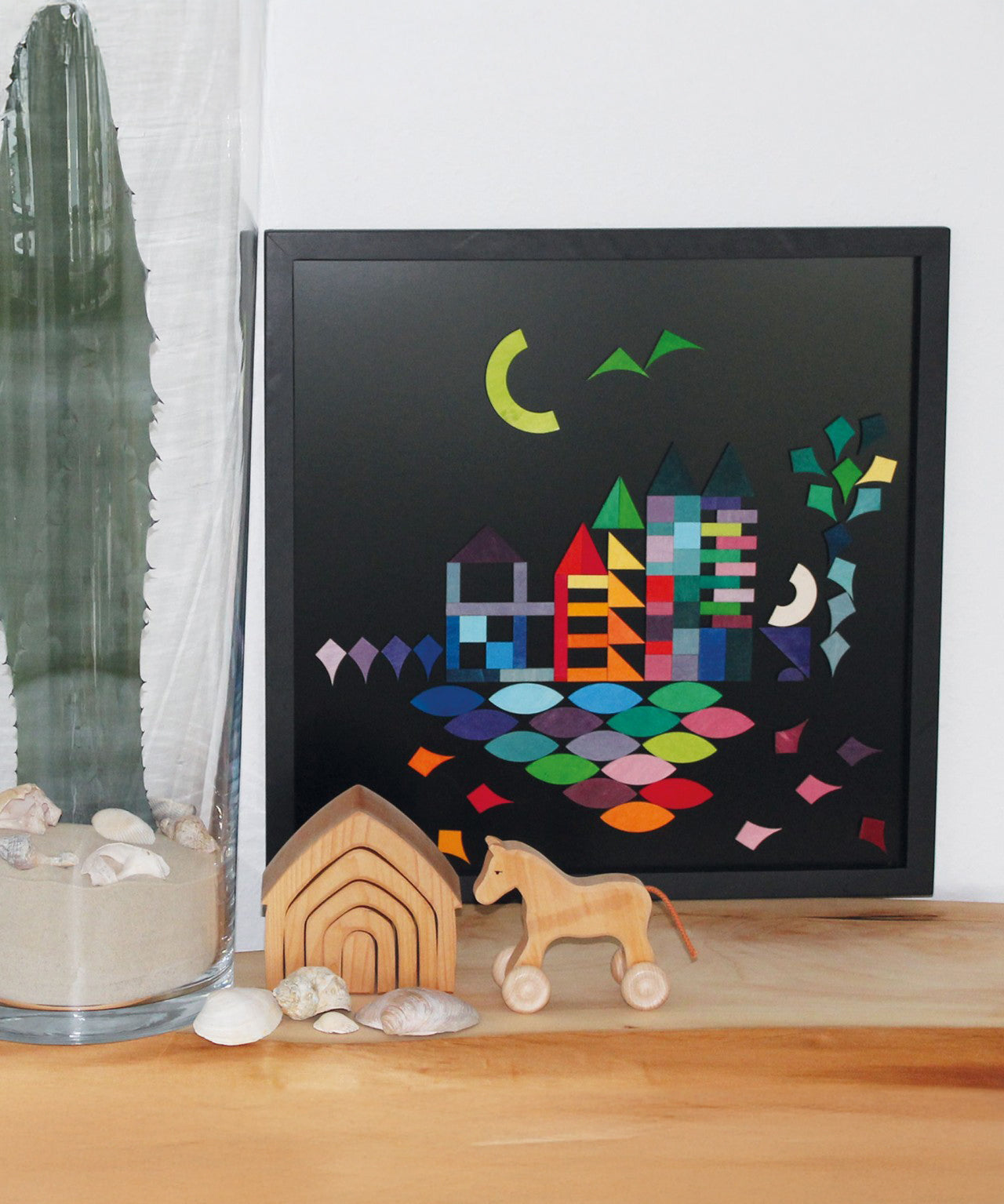 A Grimm's Large magnetic blackboard with a lovely landscape puzzle piece scene. In front of the blackboard is a small Grimm's house and horse. Next to them is a large green cactus in a glass vase