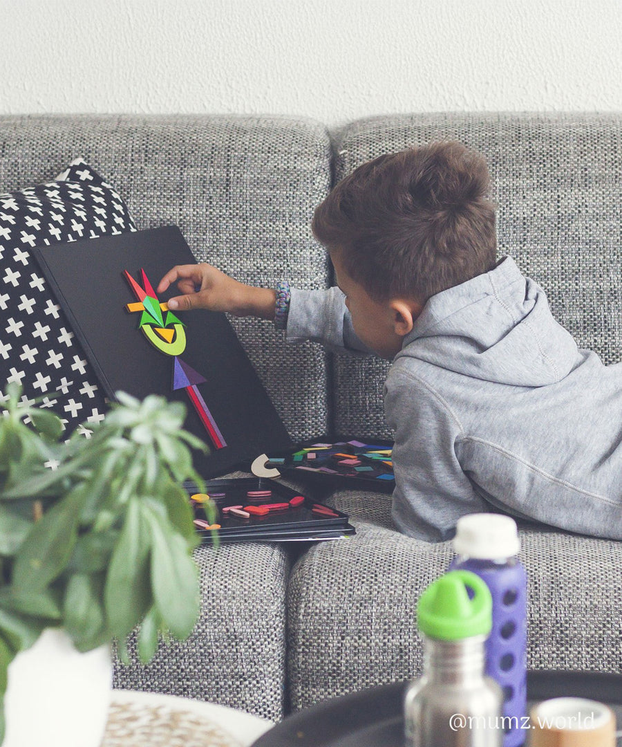 A child laying on a sofa, creating art with the Grimm's Small Magnetic Blackboard and some Grimms puzzle pieces