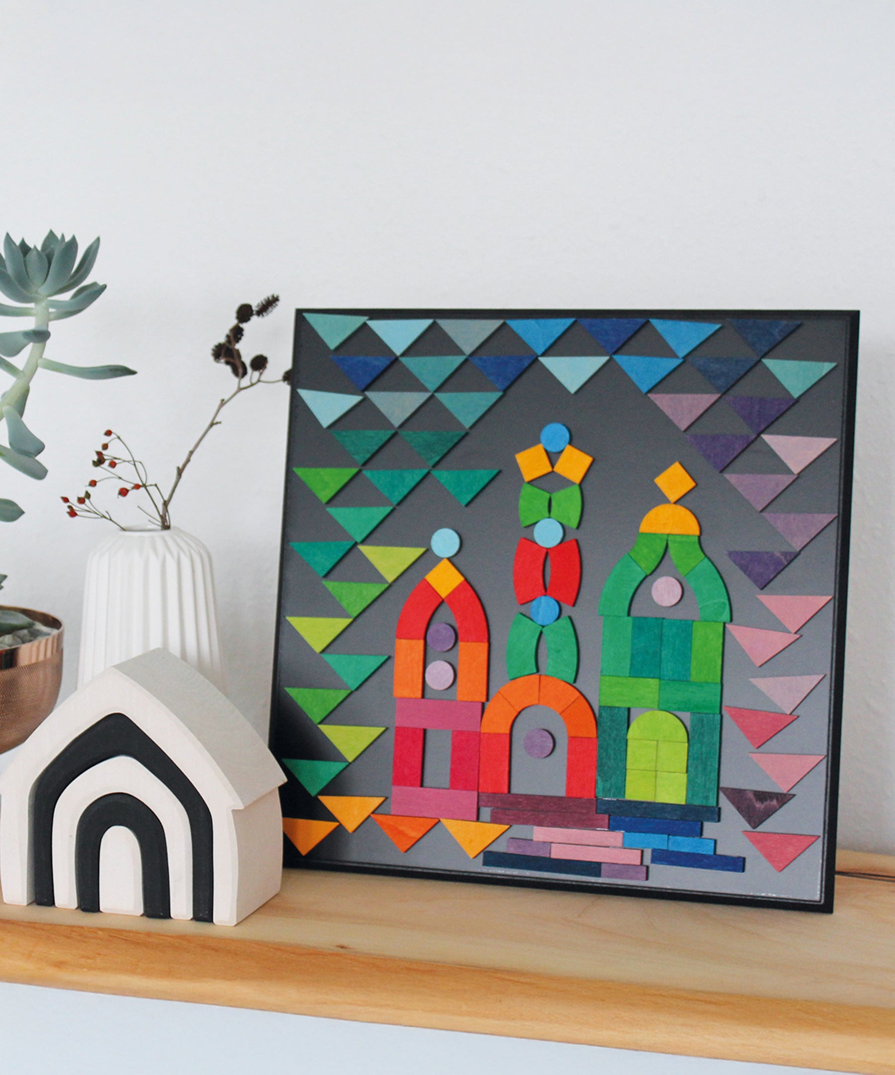 A colourful puzzle piece art work, on the Grimm's Small magnetic blackboard. In front of the board is a Grimm's monochrome house and a white vase with dried flowers in it