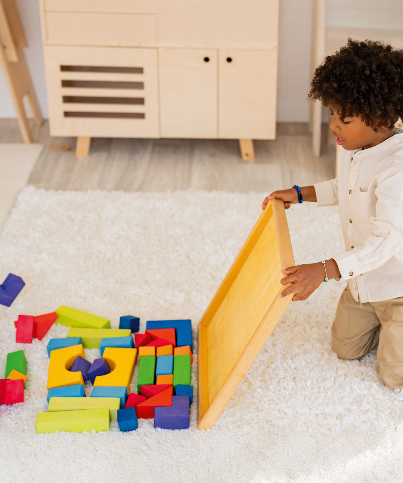 A child is sat on a fluffy cream rug, and has tipped the blocks of the Grimm's Basic Building Set from their wooden base