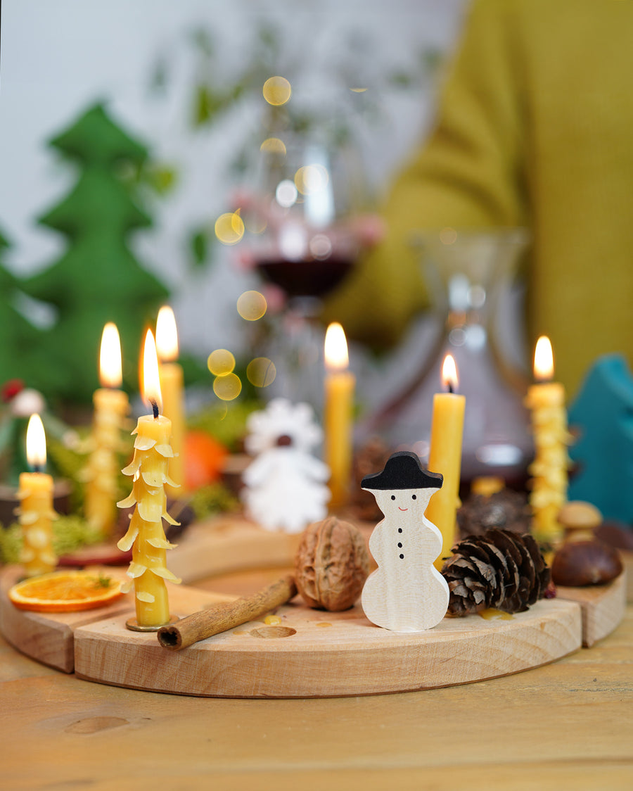 A Grimm's Wooden Celebration Ring, filled with Grimm's wooden Christmas decorative figures and candles in brass holders. There are also other decorative items such as a walnut, cinnamon stick, pine cone and conkers to create an uplifting winter display