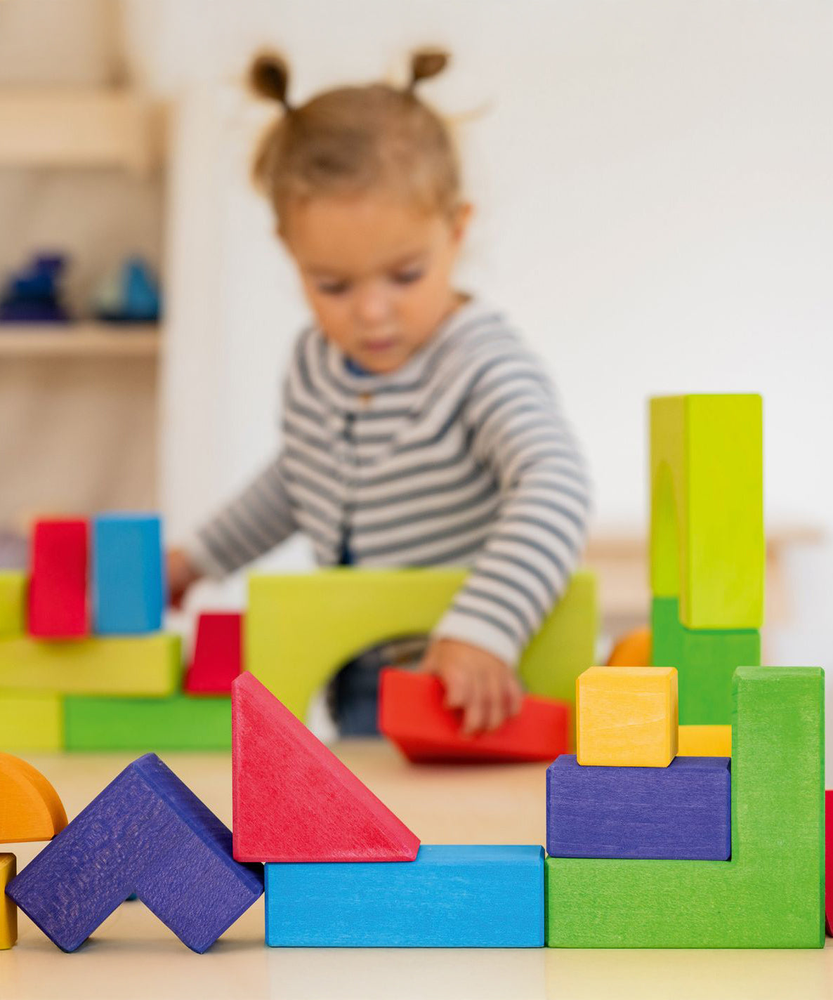 A child is happily playing with the Grimm's Basic Building Set in the back ground, with some of the Grimm's Basic Building Set Blocks in focus at the front