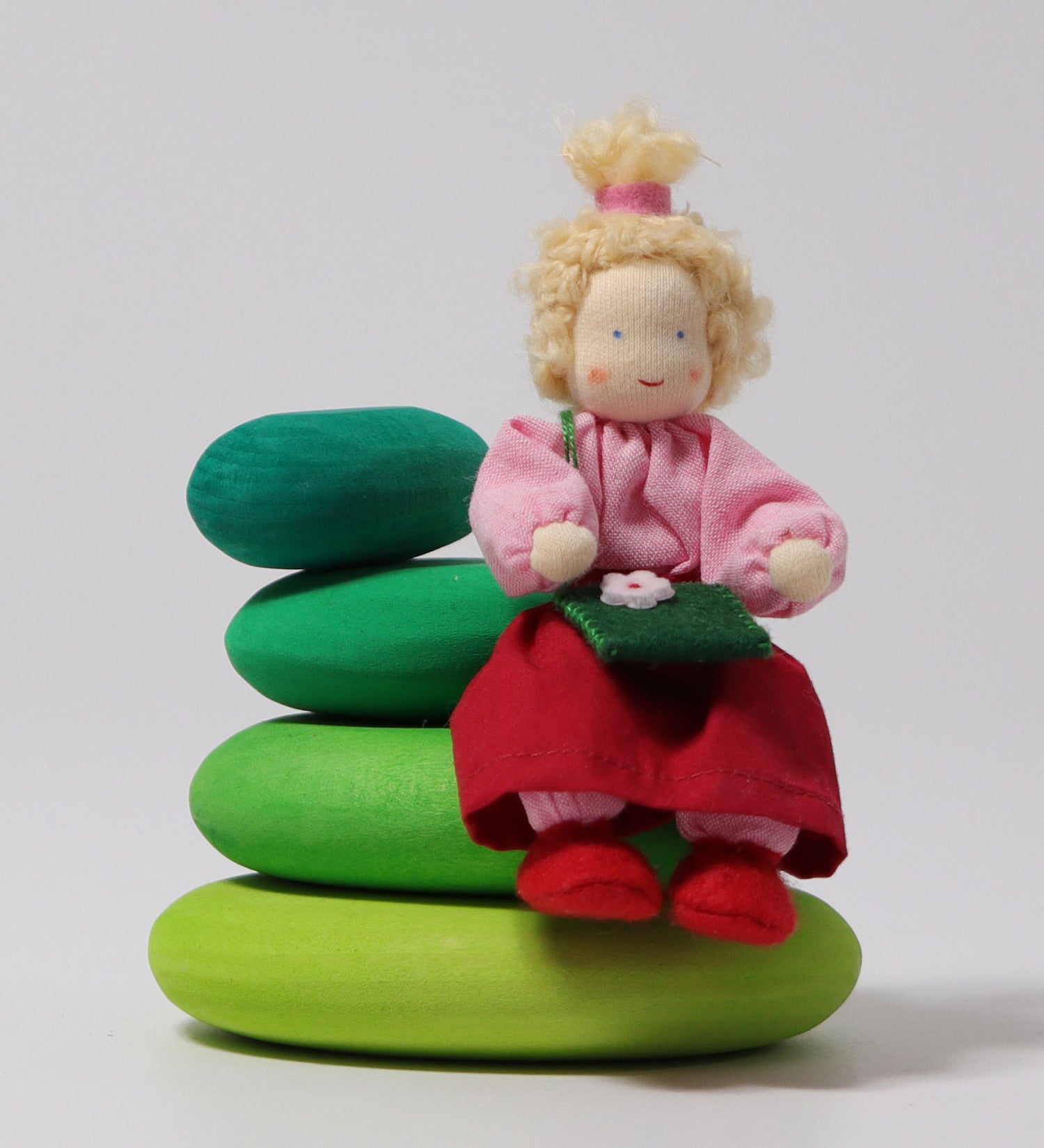 The Grimm's Blonde Girl Doll, sitting down on the Grimm's Moss Pebbles Building Set, on a grey background