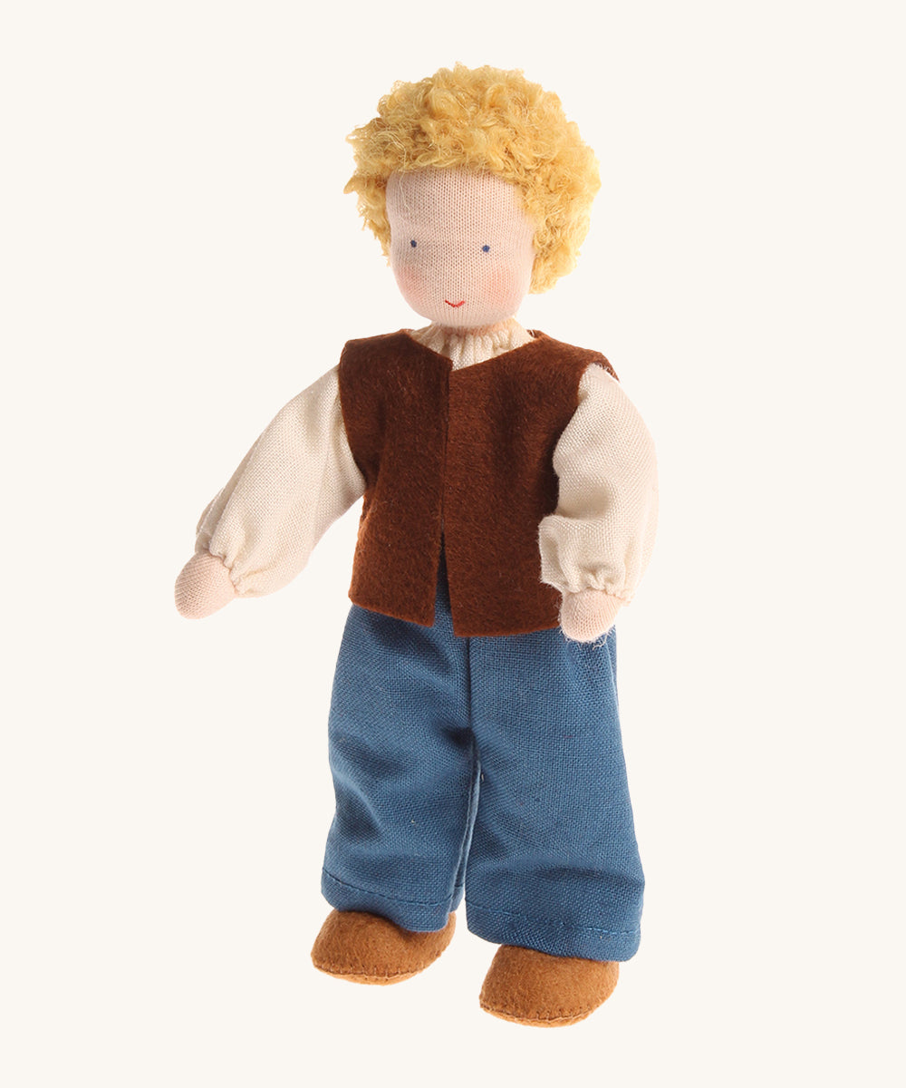 Grimm's Waldorf Blond Haired Man Doll has a soft body, blonde hair, blue eyes and is wearing light brown fabric shoes, blue trousers, a cream top and a dark brown sleeveless jacket. The image is on a cream background