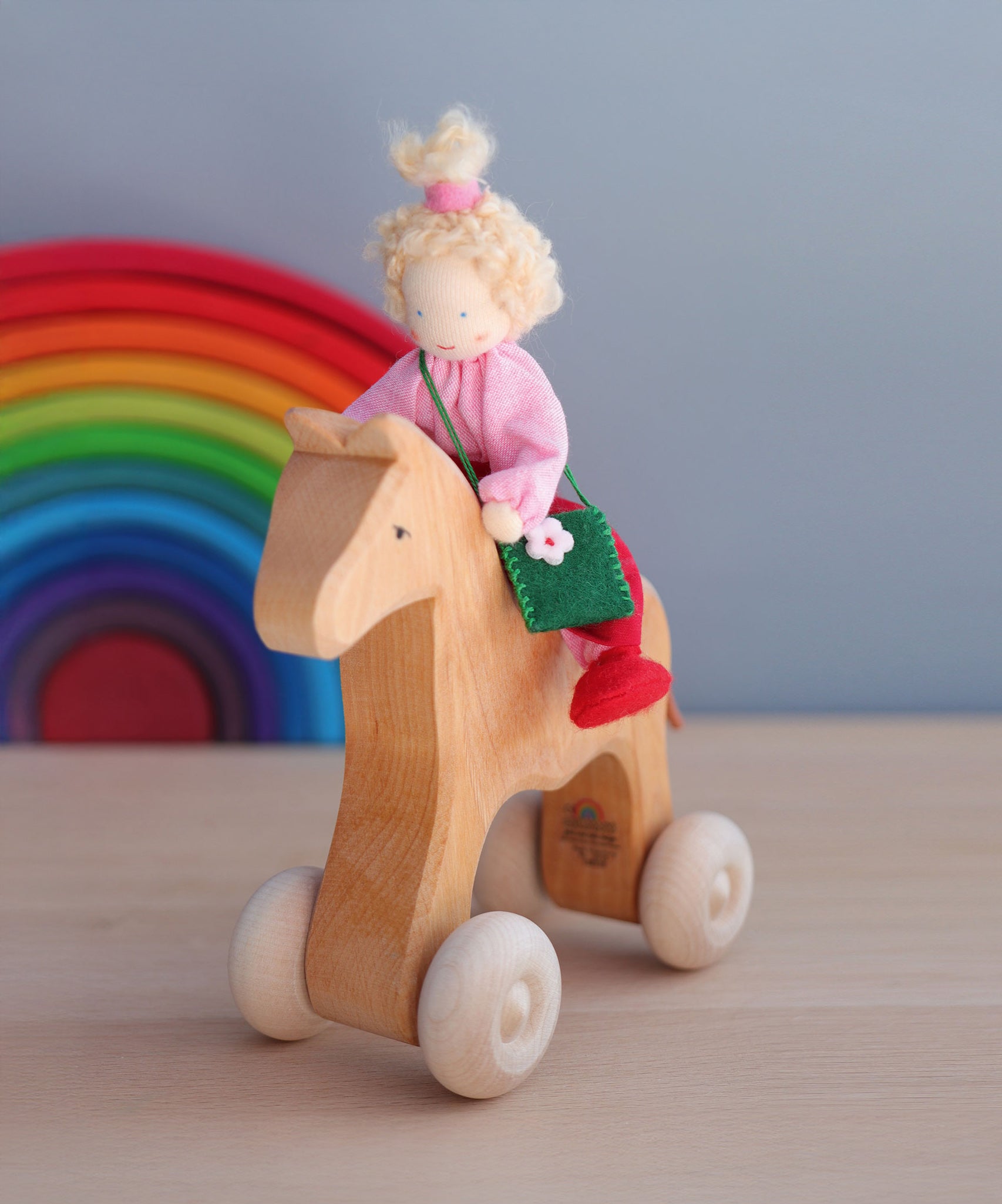 The Grimm's Blonde Girl Waldorf Doll is sat on a Grimm's Wooden Horse, with the Grimm's Rainbow 12 piece in the background