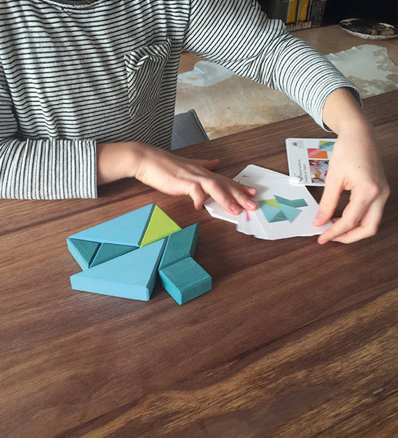 A child is playing with the Grimm's Tangram toy on a dark brown table, creating the puzzles using puzzle cards