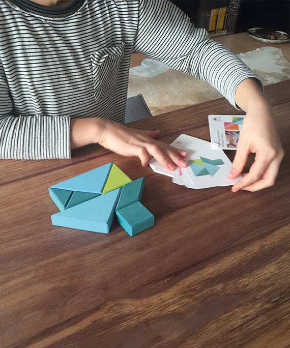 A child is playing with the Grimm's Tangram toy on a dark brown table, creating the puzzles using puzzle cards