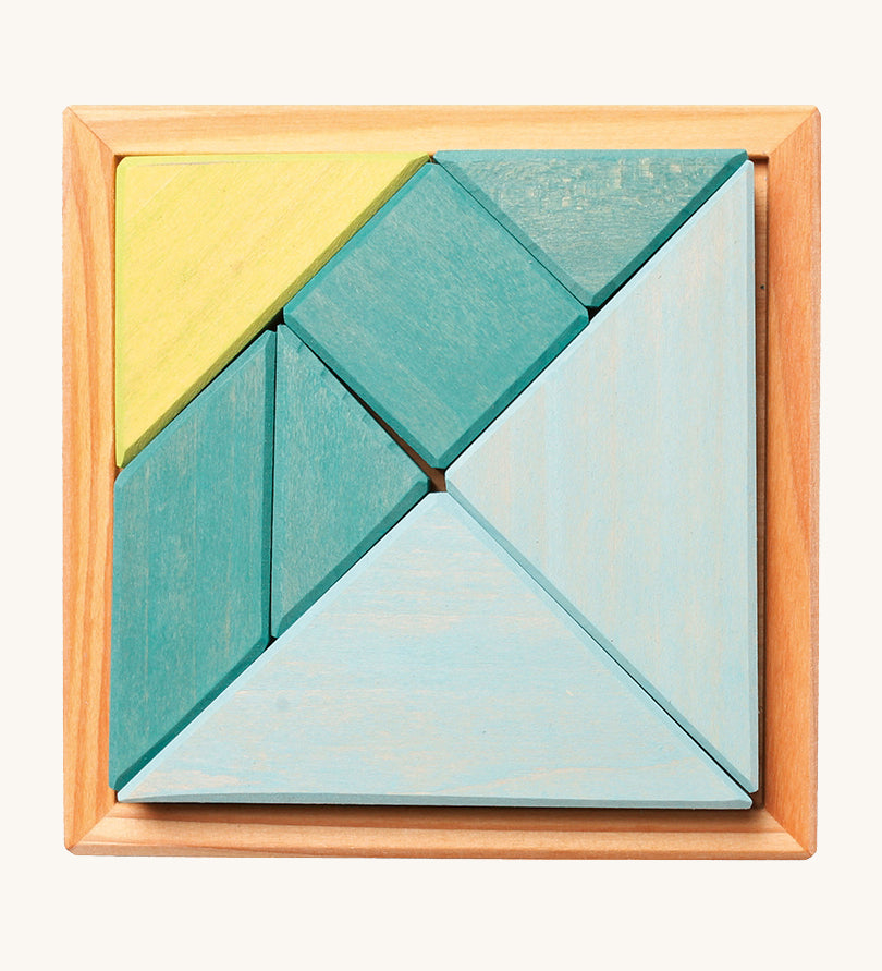 A wooden Tangram puzzle in shades of blue and green sits inside a small light wooden base on a cream background. There are 7 puzzle pieces in different shapes.