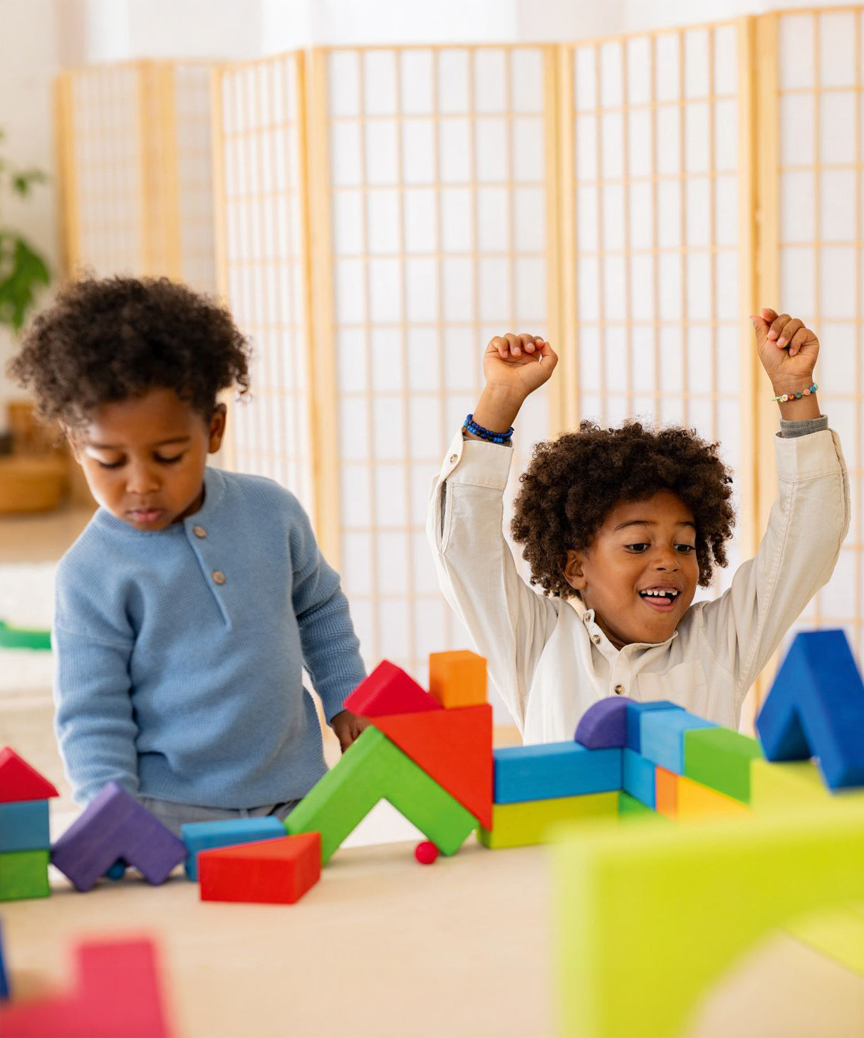 Two children are playing with the Grimm's Basic Building Set. One child has their hands up in the hair and looks excited for what they have built, and the other child is concentrating on pushing a small blue marble under a block shaped like a bridge