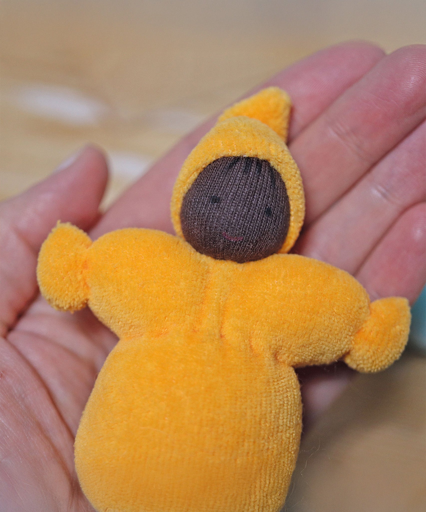 The yellow Pastel Dwarf Doll in a persons hand. A closer view of the pastel yellow Grimm's Pastel Dwarf, in a persons hand. The image shows the dwarfs body, smiling face and hat up close and gives a size guide as it fits in the palm of an adults hand.