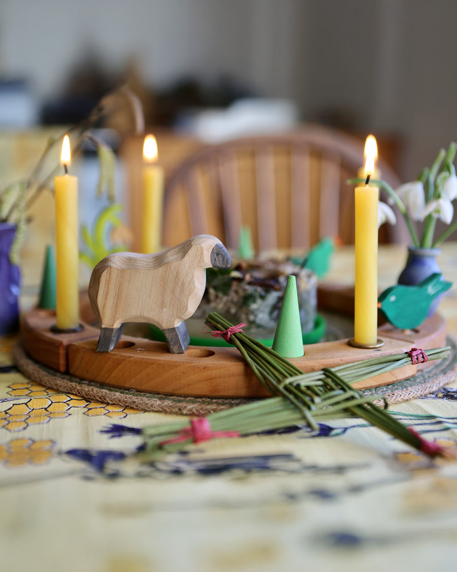 A lovely Grimm's Wooden Celebration Ring display to symbolise an Imbolc tradition. There is a sheep figure, cream coloured candles and green loose parts in the display