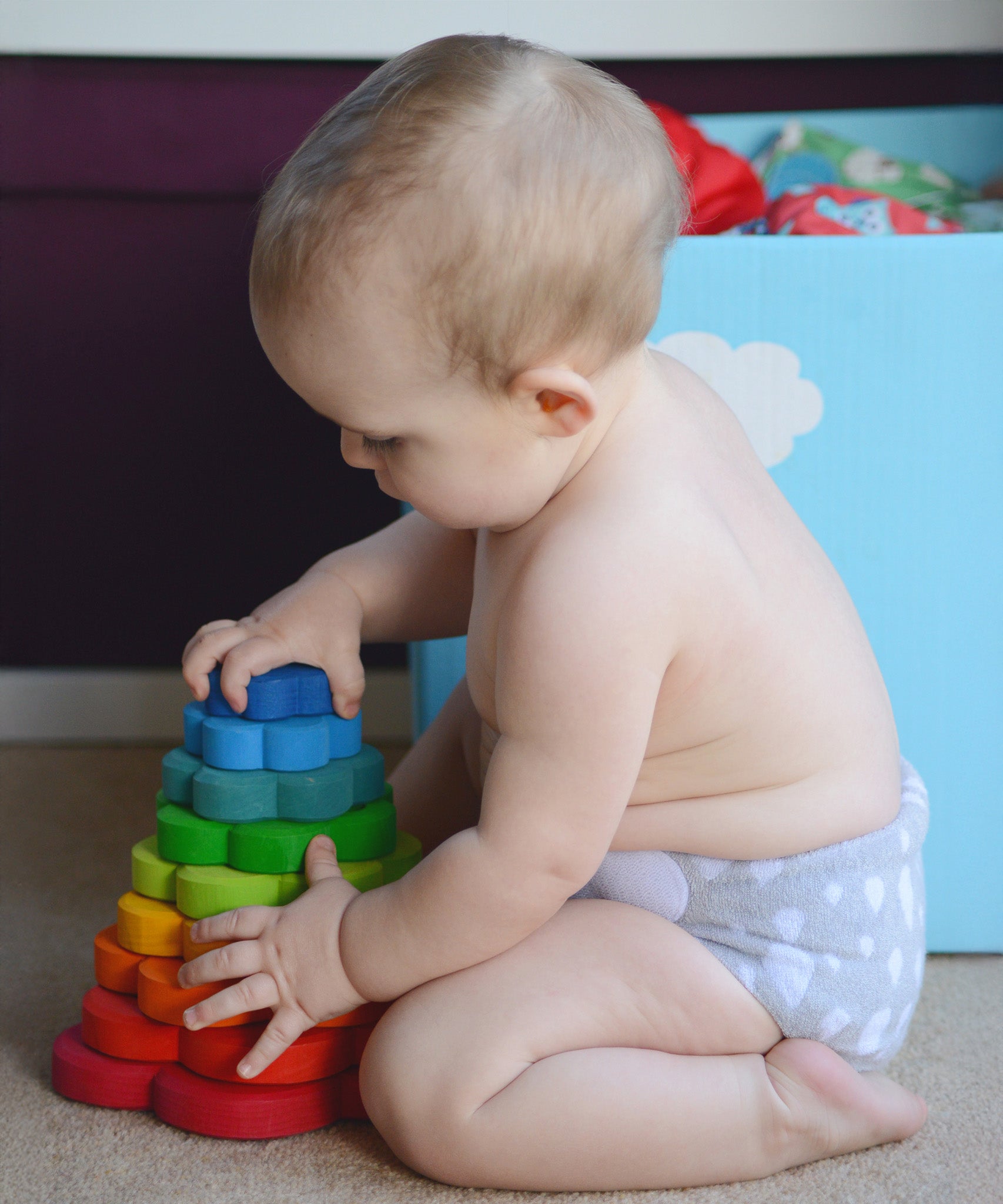 A child is sat down on the floor, infornt of a light blue toy box and is playing with the Grimm's Wooden Stacking Flower Toy. The child is holding the side of the flower and is attempting the take off the top flower block.