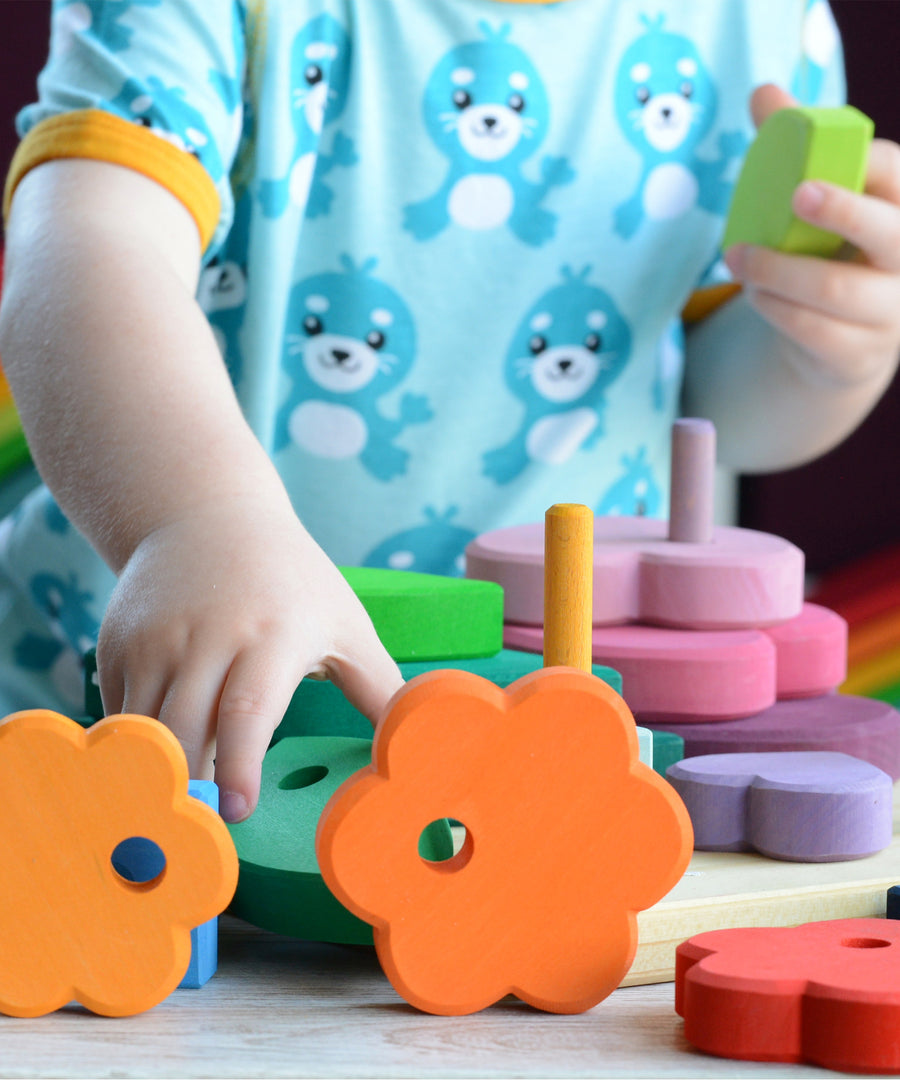 A child is playing with the Grimm's Stacking Flowers Tower. The child has taken some of the flowers off the stacker and is using them to play with