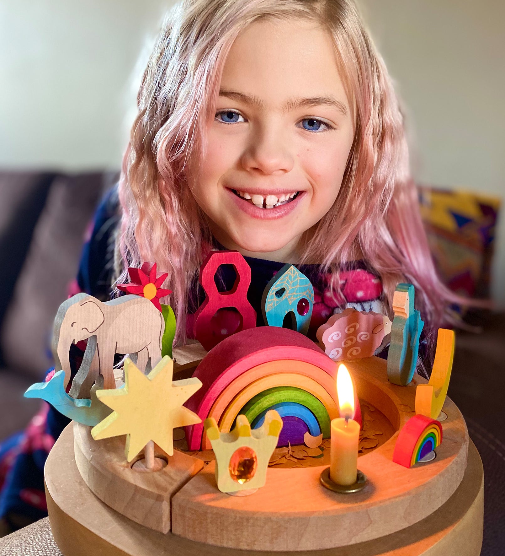 A child is celebrating their 8th birthday and has in front of them, a Grimm's Waldorf celebration ring filled with Grimm's decorative figures, a candle and a Grimm's number 8. In the centre is a Grimm's 6 piece mini wooden rainbow.