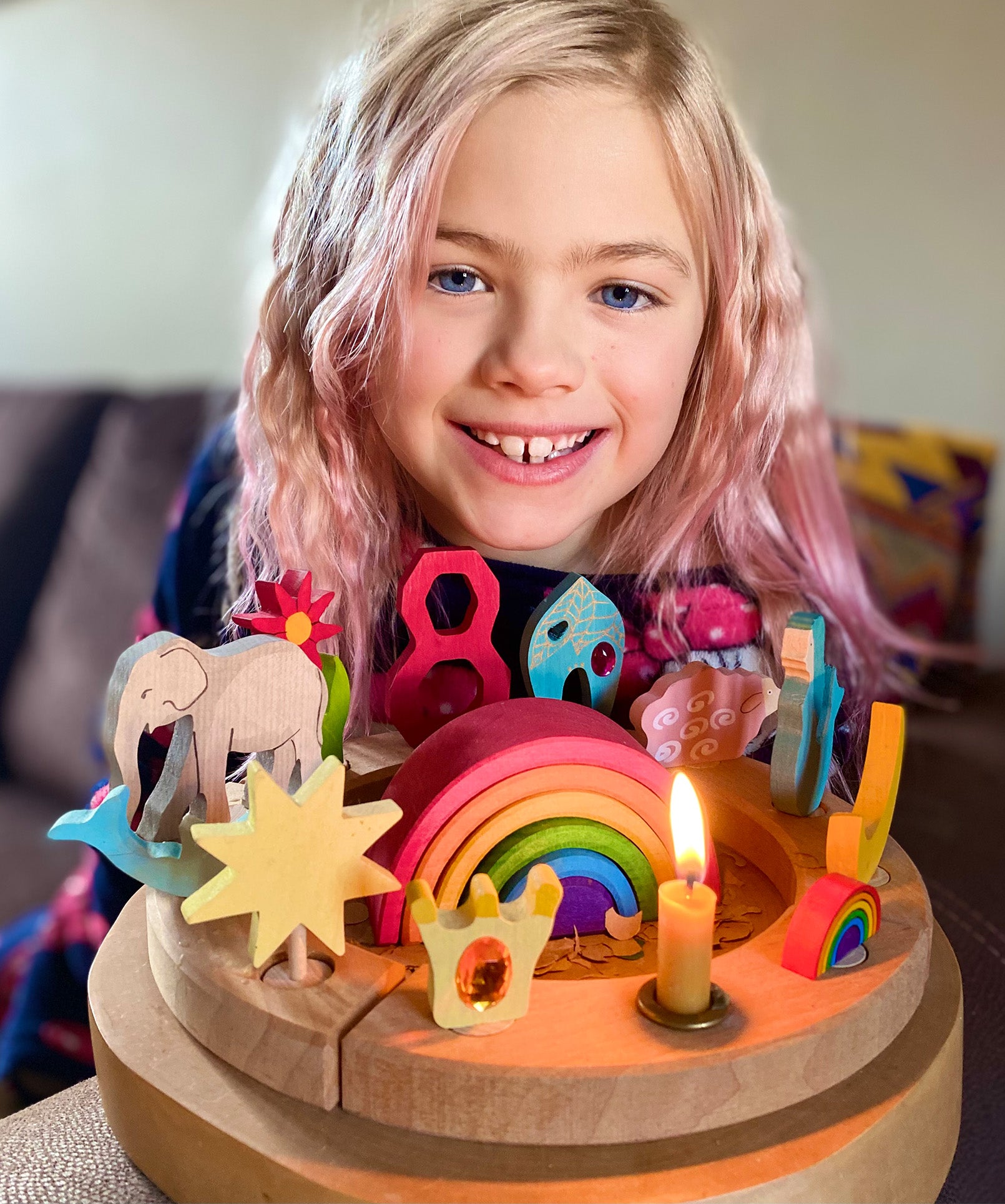 A child is celebrating their 8th birthday and has in front of them, a Grimm's Waldorf celebration ring filled with Grimm's decorative figures, a candle and a Grimm's number 8. In the centre is a Grimm's 6 piece mini wooden rainbow.