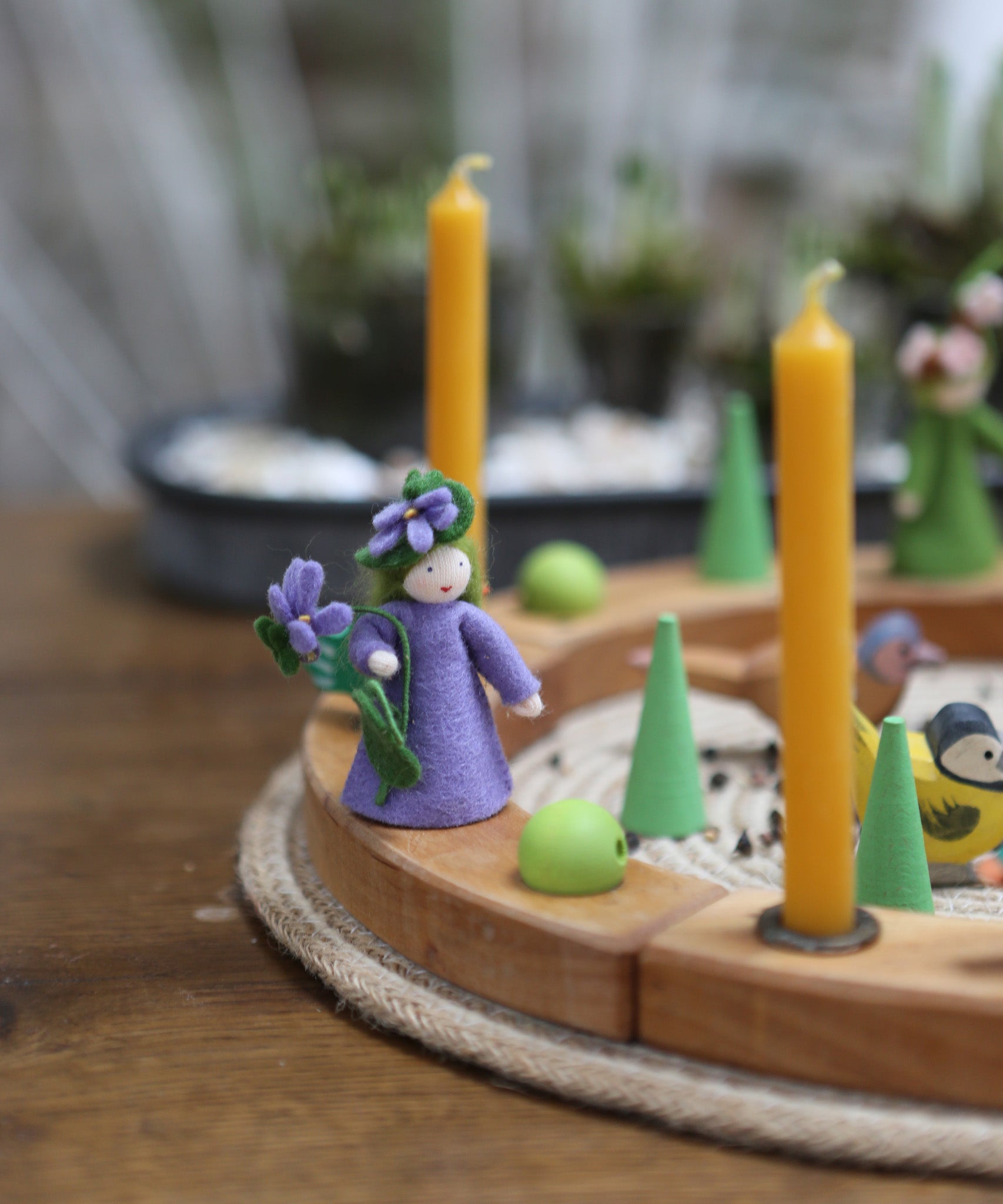 A beautiful Ambrosius doll sits on a Grimm's Wooden celebration ring, with yellow candles and green loose parts, to create a vibrant spring celebration display