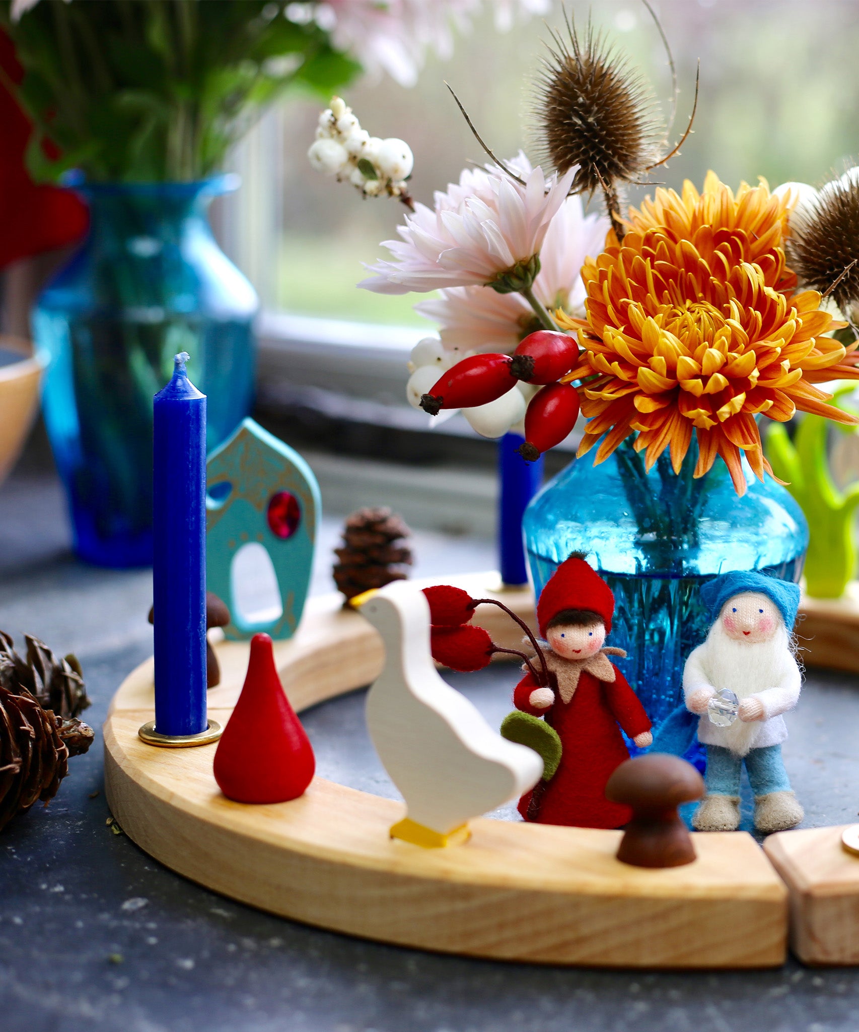 A Grimm's Wooden Celebration ring is used in a pretty winter display using Grimm's candles, Grimm's decorative figures and other loose parts. In the centre is a blue vase with flowers, and two Ambrosius dolls