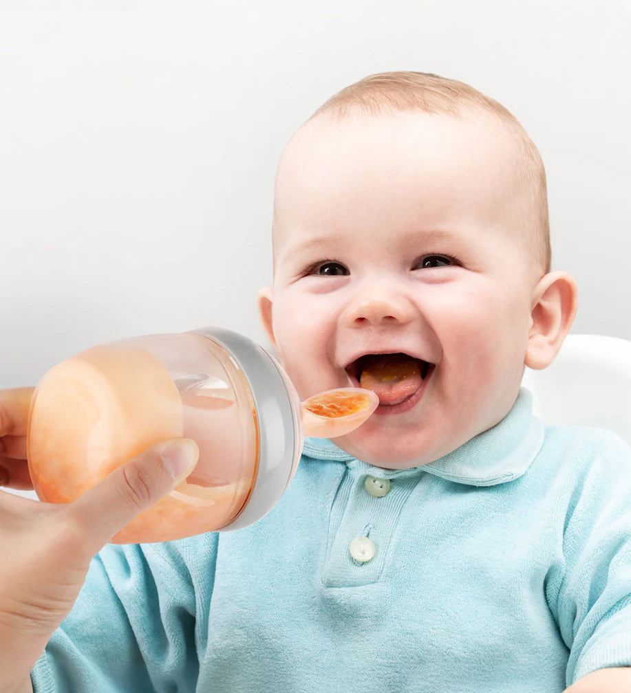 A baby smiling and eating from the Generation 3 Silicone Bottle Feeding Spoon Head, as their parent holds the feeding spoon to the babies mouth