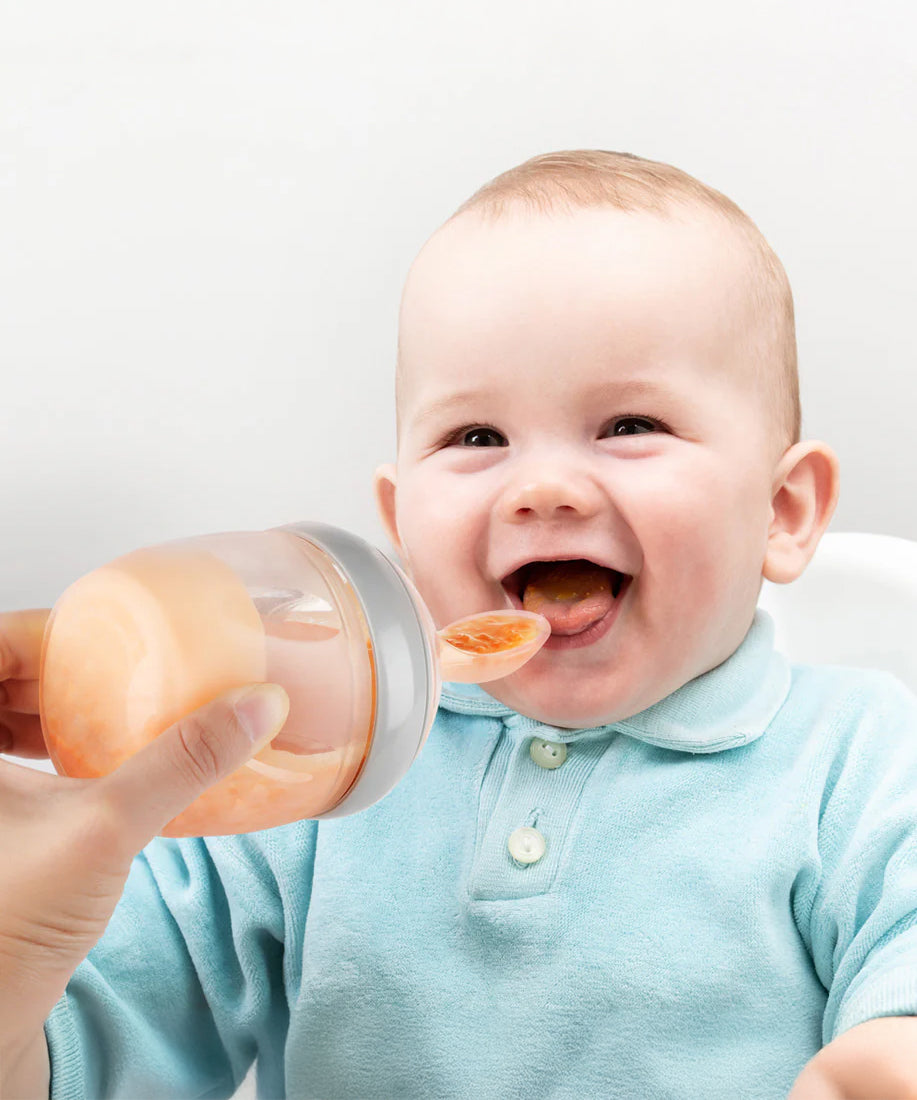 A baby smiling and eating from the Generation 3 Silicone Bottle Feeding Spoon Head, as their parent holds the feeding spoon to the babies mouth