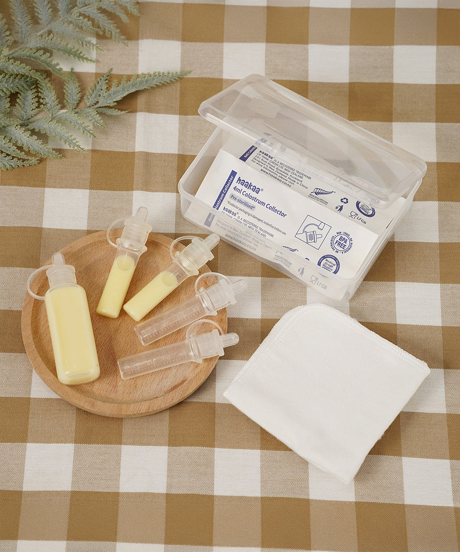 The Haakaa Silicone Colostrum Collector multi-size pack is sat on a  small light brown wooden dish, with a small open box with Silicone Colostrum Collectors inside in their packaging. Underneath is a table cloth in cream and brown check print