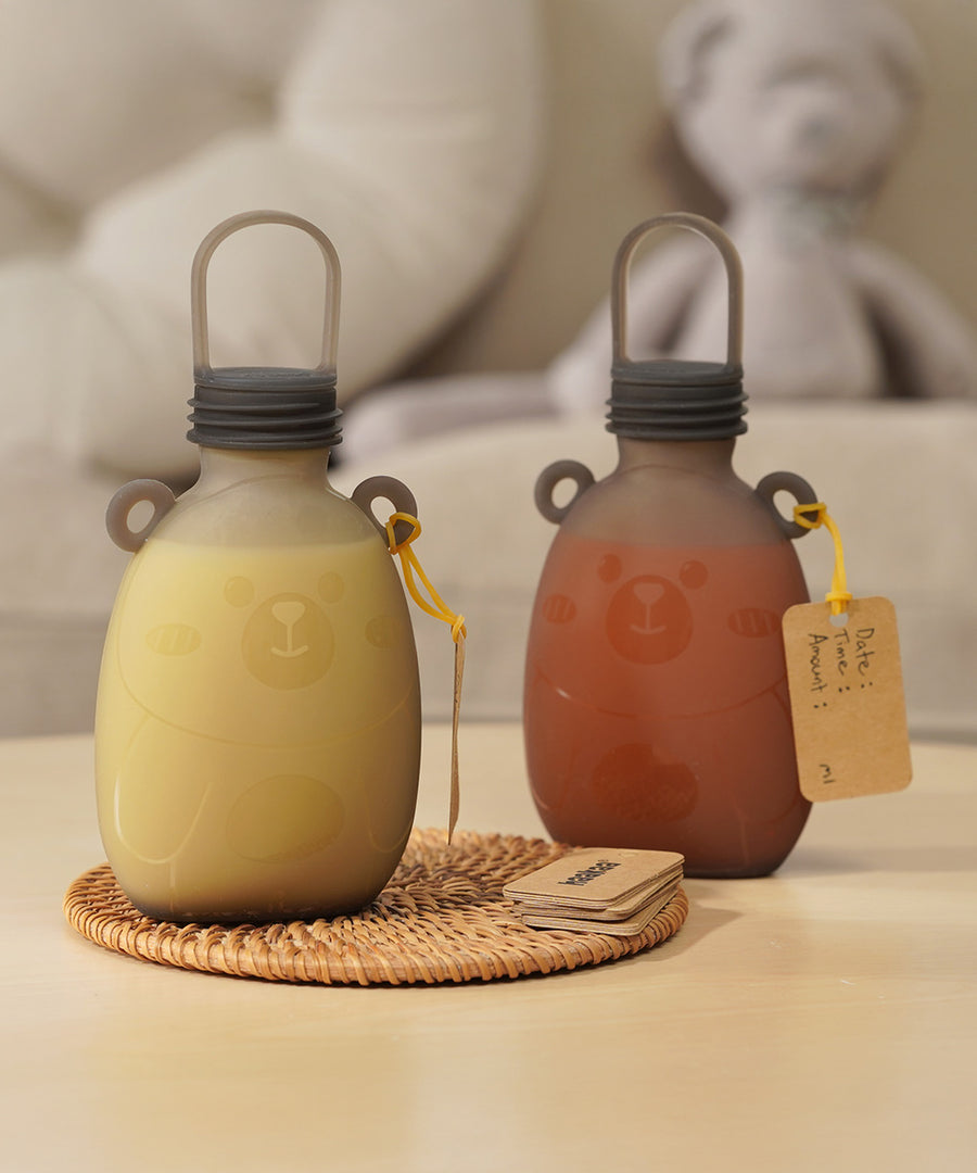 Two Haakaa Happii Bear Silicone Goody Pouches on a table with a label attached to the bear ear on the bottle. The left pouch is filled with yellow liquid, and the right is filled with orange/red liquid