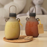 Two Haakaa Happii Bear Silicone Goody Pouches on a table with a label attached to the bear ear on the bottle. The left pouch is filled with yellow liquid, and the right is filled with orange/red liquid