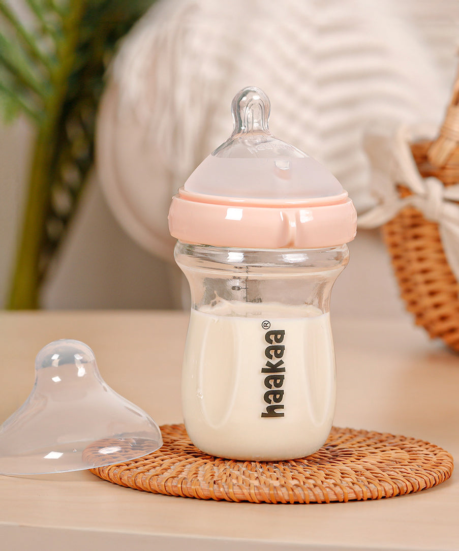 The Haakaa Generation 3 Glass Baby Bottle in peach, stood on a wooden mat, with the bottle lid next to it