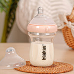 The Haakaa Generation 3 Glass Baby Bottle in peach, stood on a wooden mat, with the bottle lid next to it