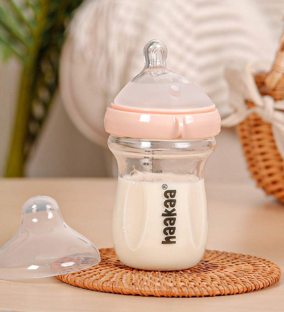 The Haakaa Generation 3 Glass Baby Bottle in peach, stood on a wooden mat, with the bottle lid next to it