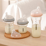 The Haakaa Generation 3 Glass Baby Bottles in 120ml /180ml/300ml, stood on a light brown table with a cork mat under 2 bottles