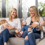 Two people sat in a living room, breast feeding their children with one holding the Generation 3 160ml Breast Pump in peach